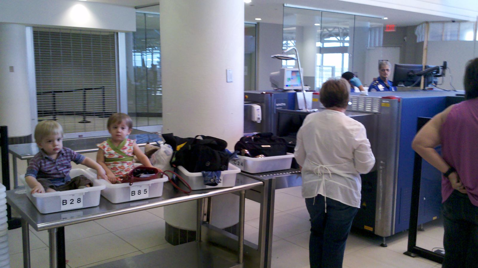 Boy-girl twin toddlers in TSA security bins, about to go through x-ray machine. TSA does not look happy.